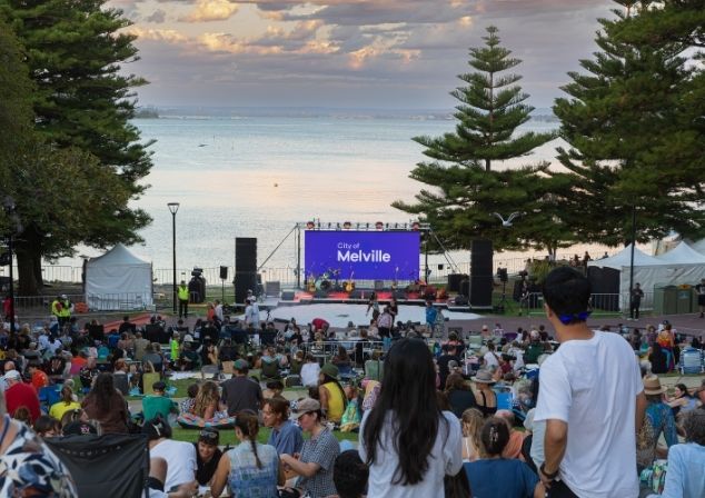 Audience members at an event, sitting on picnic blankets, a stage set up by the river. 