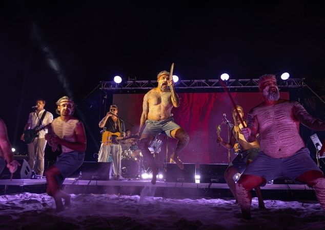 Audience members at an event under the pine trees, some standing, some sitting.