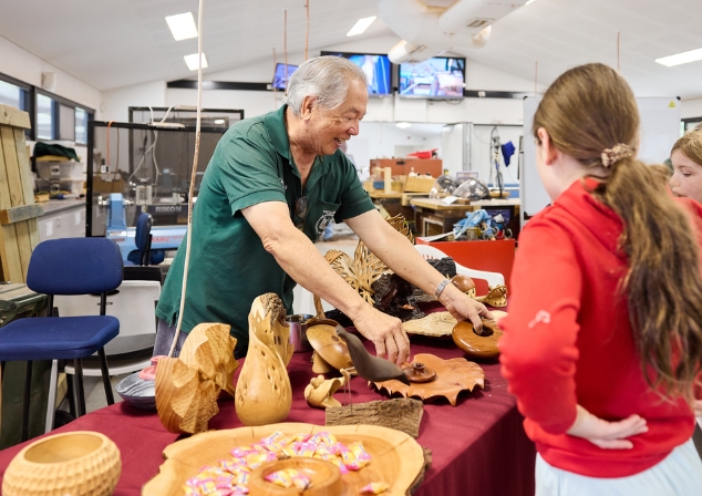 Gentlemen standing behind a table of woodworked pieces smiling at two young girls looking at his work.
