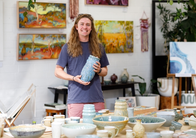Young man with long ginger hair smiling at the camera, holding a tall blue ceramic hand made vessel, standing behind a table of hand made ceramic pieces in a gallery space.