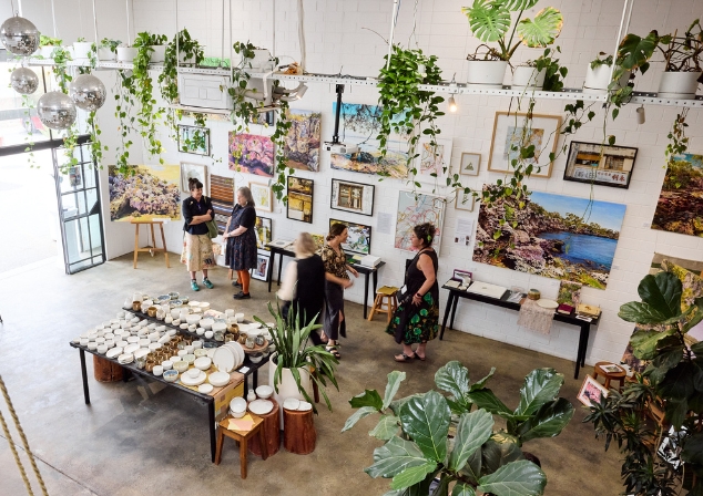 Inside of the Feld Space with art on walls, plants hanging down, table of ceramics in the centre and people standing round talking.