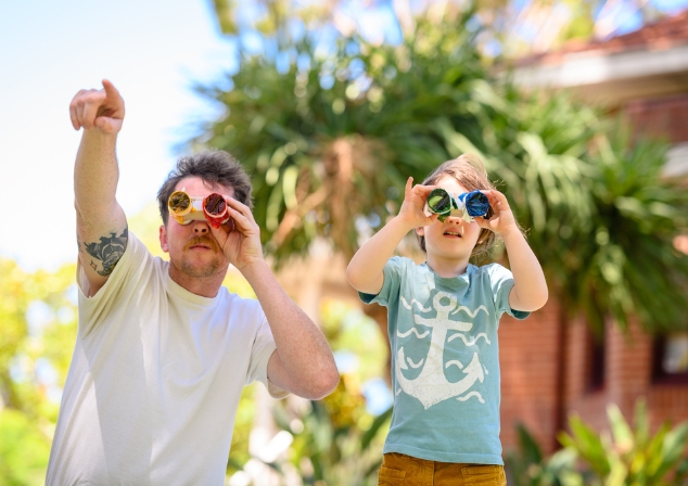 A man and a child using homemade binoculars with colourful cellophane looking into the sky, the man is pointing at something in the distance.
