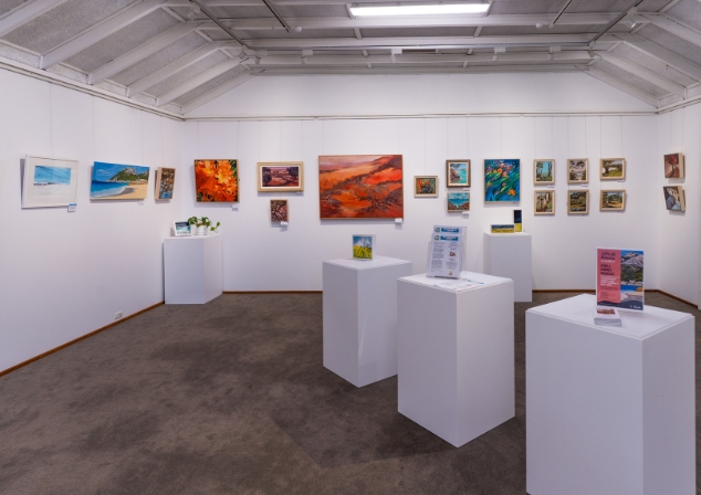 Atwell Gallery exhibition room with paintings on the wall and plinths set up in middle of room with artworks and brochures.