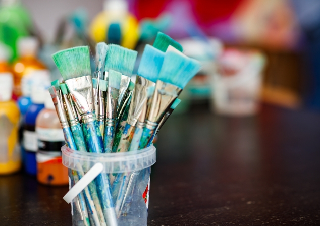 Plastic container filled with blue stained paint brushes, brightly coloured paints blurred in the background.