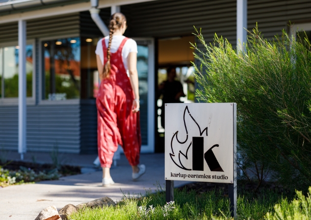 The front of a building with sign Karlup Ceramics Studio, girl in bright red overalls is walking into the building.