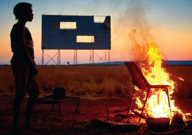 First Nations boy standing outside at twilight nearby to a campfire - still picture taken from the promotional poster for the film Satellite Boy