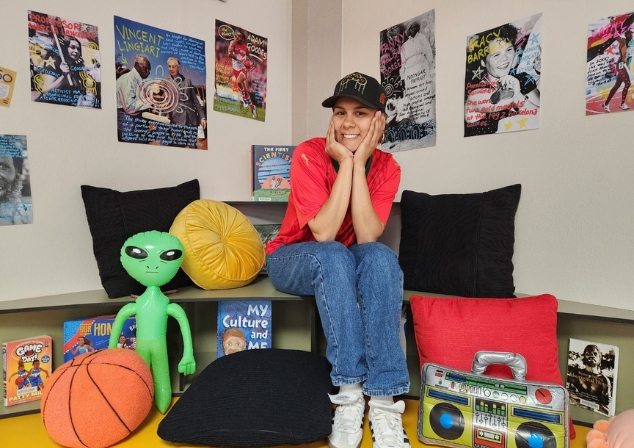 Ilona McGuire sitting with hands in face smiling in front of a library installation of Aboriginal authors and inspiring posters with Aboriginal sports people and important historical figures.