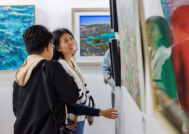 Two people standing in an art gallery, one woman is staring happily at an artwork that we can't see and her face is reflected back from the glass of another artwork. 