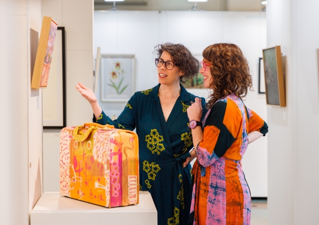 Two woman in patterned and brightly coloured dresses, wearing glasses, looking joyously at a painting with a brightly coloured painted suitcase in the foreground. 