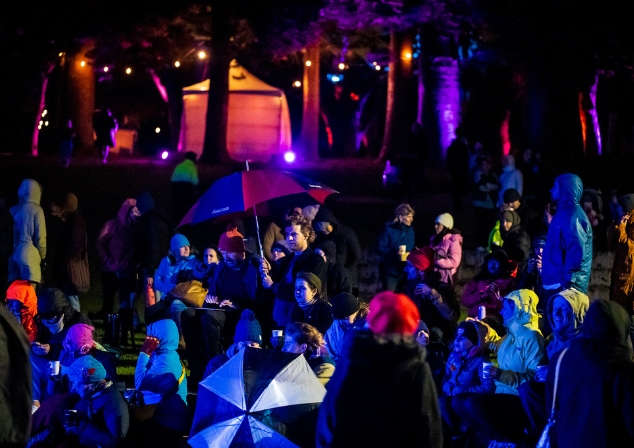 Crowd of people in rain jackets, with umbrellas, outdoors at night with a performance tent and lights in the background.