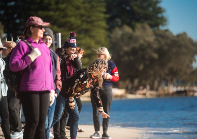 People lined along the river bank, one woman has a hand extended throwing sand into the river.