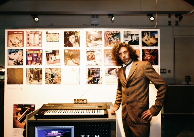 Man in brown suit next to keyboard with a wall of records in the background as part of Cold War to Dance Floor exhibition.
