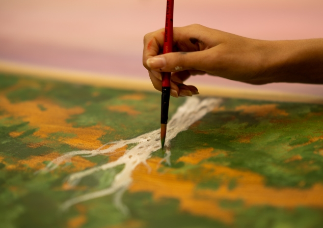 A hand holding a paintbrush with green paint adding detail to a landscape painting.
