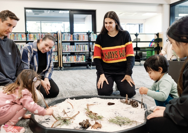 Cultural educator Rickeeta Walley leading a storytime session with children and families sitting around a container with sand and natural materials.