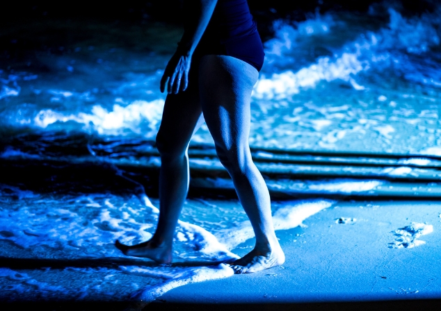 It's night-time, a swimmer in black one-piece bathing suit is seen from the waist down, walking into a the shallows of the river from the river beach. 