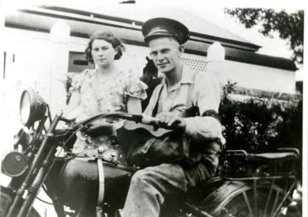 Black and white photograph of a man (Clem Booth) sitting on a motorcycle and woman (Rose Shorthouse) standing beside him. A house can be seen behind.