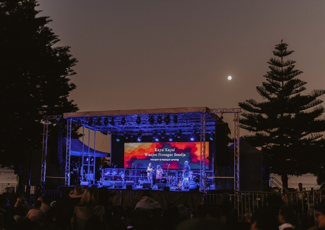 A stage lit up in twilight on the shores of Dyoondalup Point Walter with a moon behind and audience seated in chairs in front. 