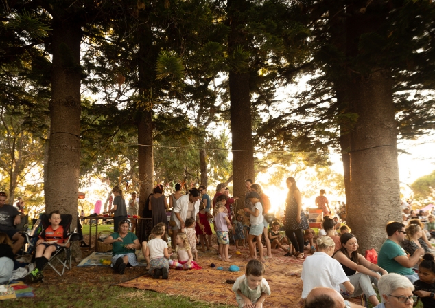 Audience members at an event under the pine trees, some standing, some sitting.