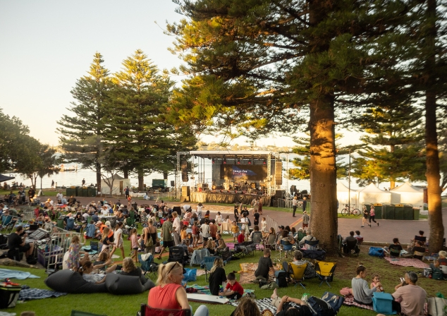Audience members at an event, sitting on picnic blankets, a stage set up by the river. 
