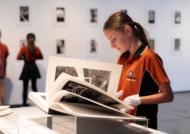 A primary school aged student looking at a large book of lino-prints, wearing white gloves, in a gallery.