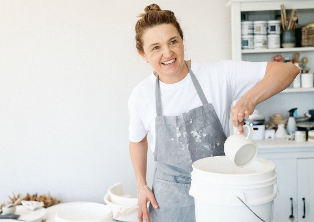 Women in white tshirt and grey apron dipping a pottery cup into a bucket of glaze, smiling looking off to the right of the picture.