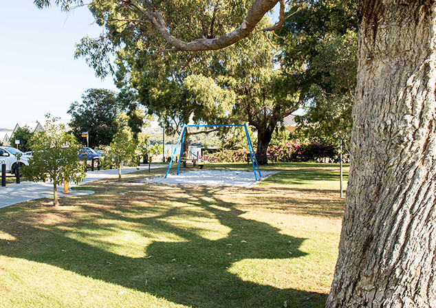 Photo of grass area with a swing set in a sandpit in the centre. Footpath and cars to the left, trees around area.