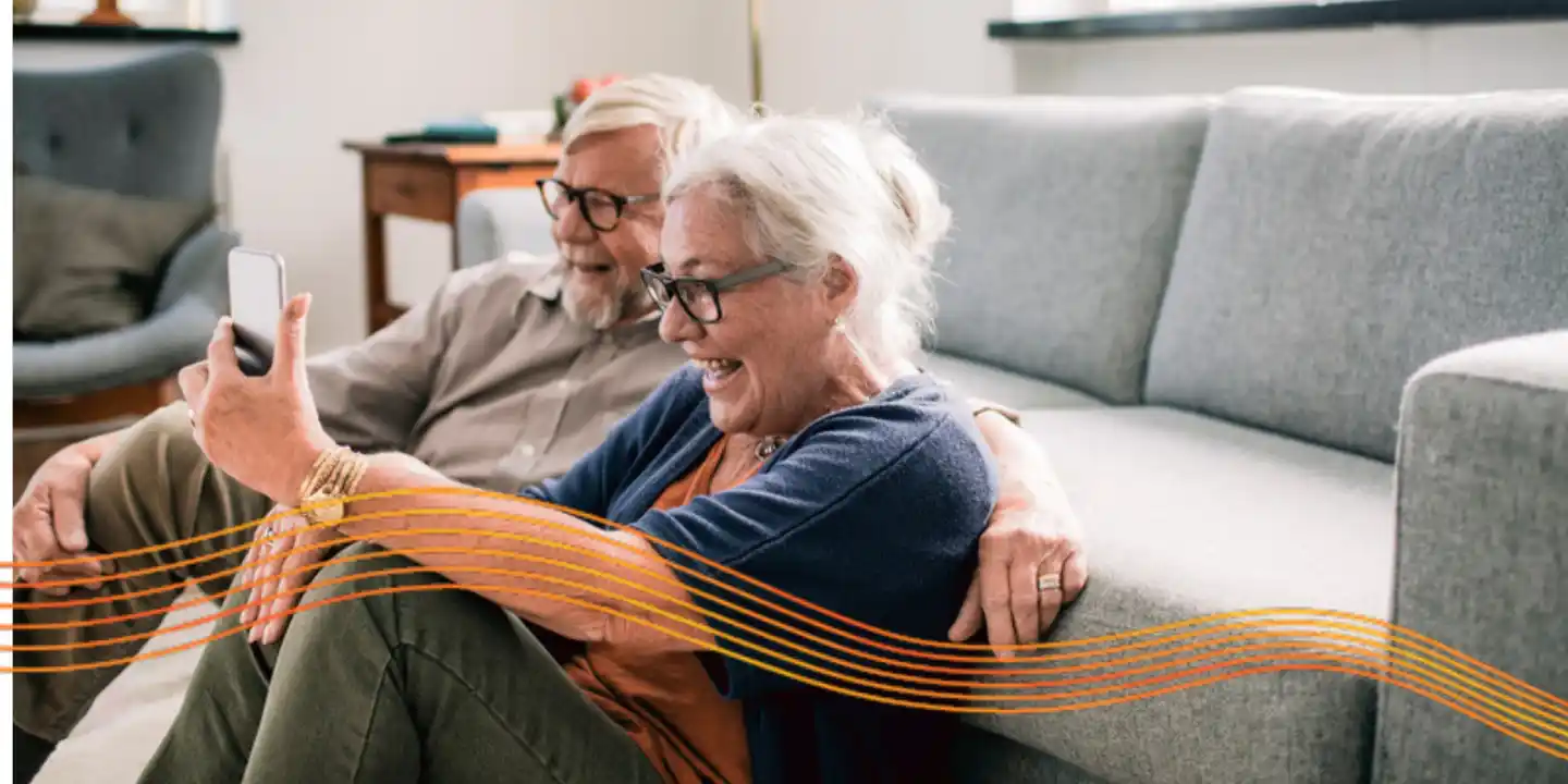 An older couple sit on the floor in front of a couch smiling. The woman is holding a smartphone, looking into its screen.