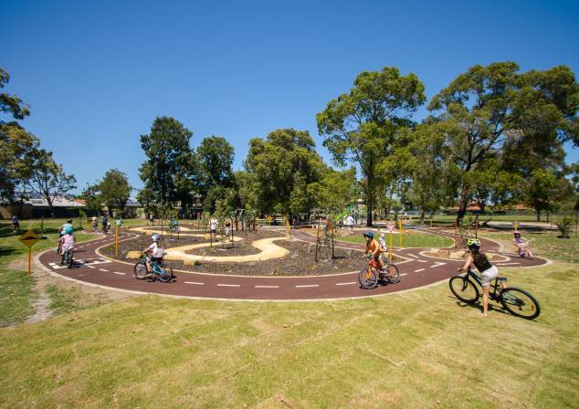 Photo of lawn area with a bike track in the middle. Large group of children riding bikes on track.