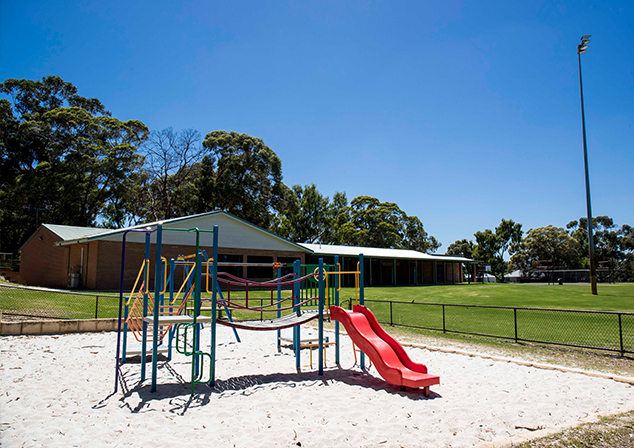 Children's playground in sand pit with tall trees surrounding with grass and building behind