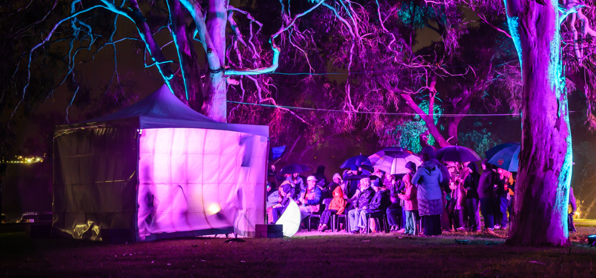 Audience huddled under trees, lit up by purple light watching a performance at Melville Midwinter