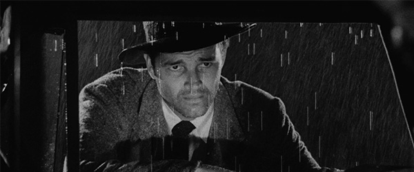 A black and white image of a man wearing a fedora, leaning in a car window.
