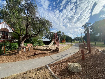 Image of footpath going through mulch area with timber playground equipment installed on it. Bright cloudy sky in background.