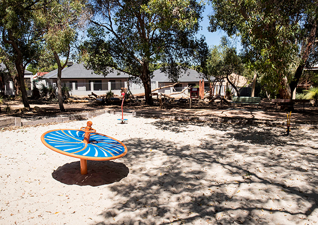 Picture of sand pit with various playground equipment. Trees and houses in background.