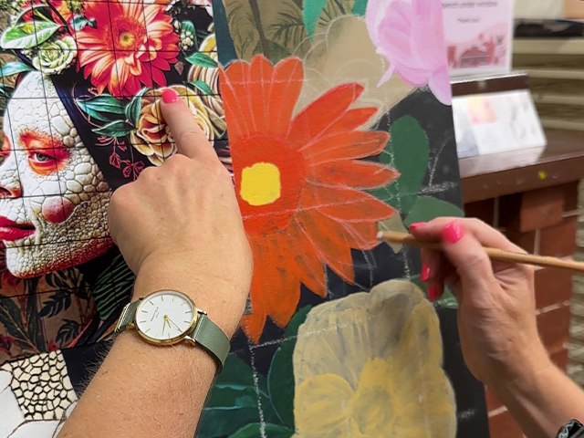 A hand paints a canvas with acrylic paint and a paintbrush. The painting subject is a bright red flower.