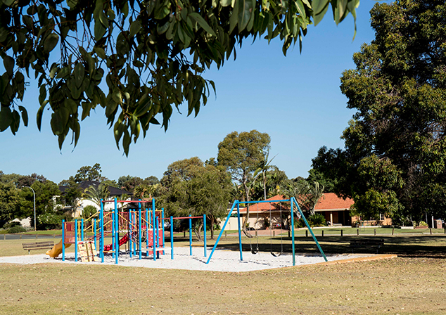 Image of grass field with playground in a sandpit placed in the middle. Trees around area, leaves hang in foreground.