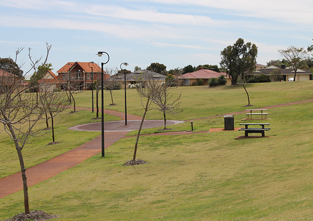 Photo of grass field with a footpath cutting through on left. Skinny, bare trees and tall light posts decorate the sides of the path. To the right are park benches and a bin. Houses in background.
