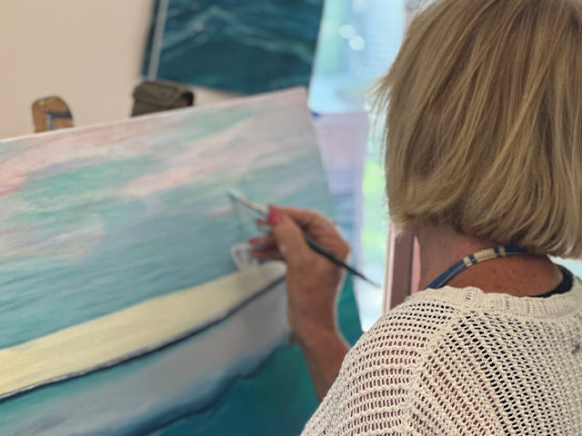 A woman painting a canvas with acrylics, the subject is a soft blue sky, white clouds and a soft yellow sand beach.