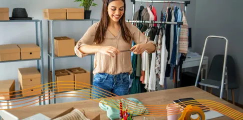 A woman taking a photo of an item of clothing laid out on a table. In the background there are boxes on shelves and clothing on a rack.