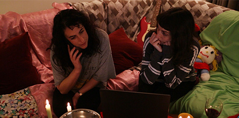Two teenage girls sit in a candle lit room in front of a laptop. One of them is on the phone.