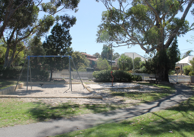Photo of grass area with large trees and a footpath cutting through. A sandpit with a swing set and other playground equipment in the middle of area.