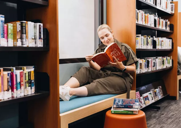 A woman sits on a bench between library shelves reading a novel.