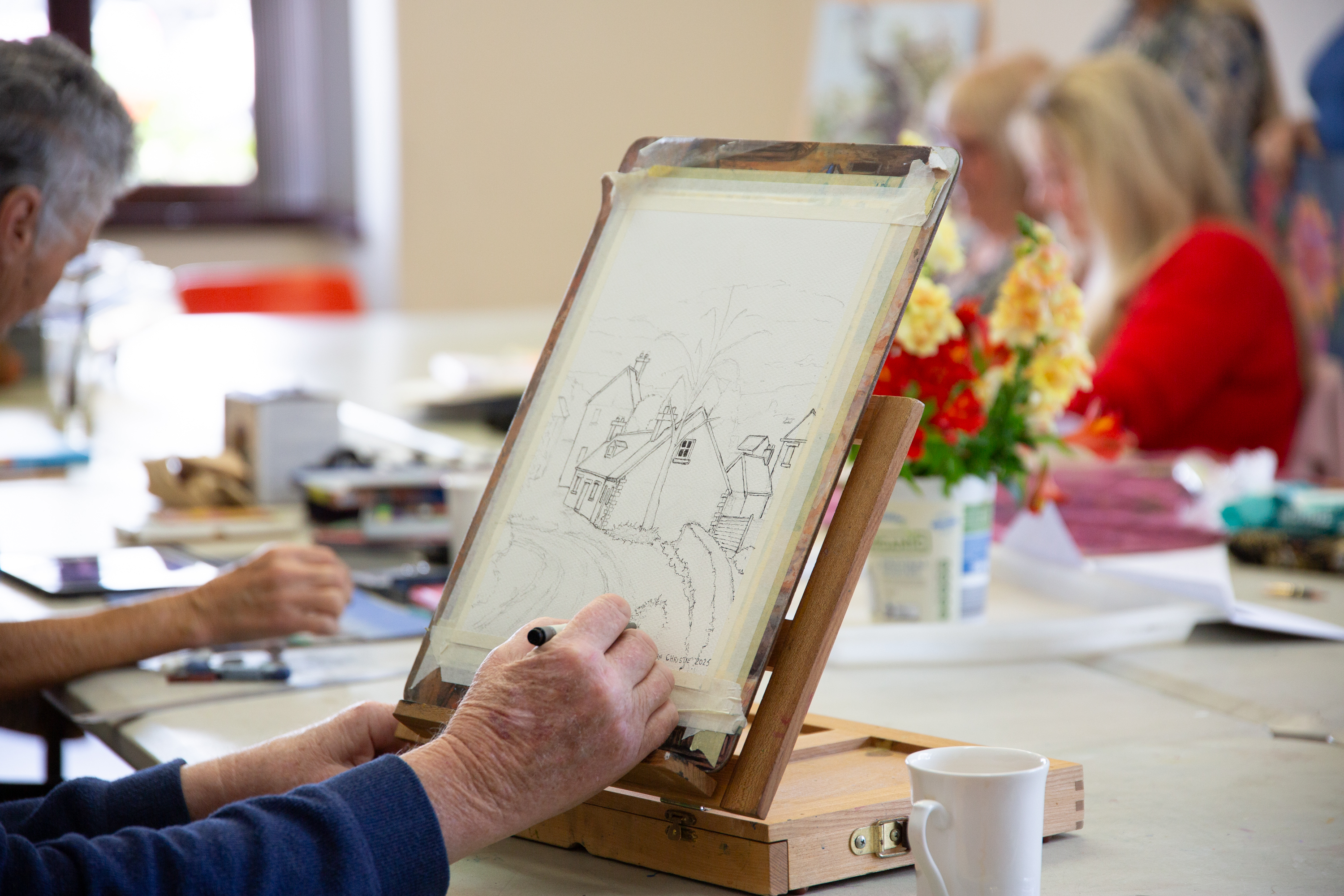 A hand drawing with a pencil on a small canvas. In the background, others work on their own artworks. There is a bouquet of flowers in the middle of the table.