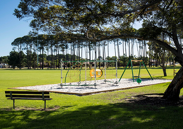 Photo of playground in grassy area. Park bench in foreground, trees in background.