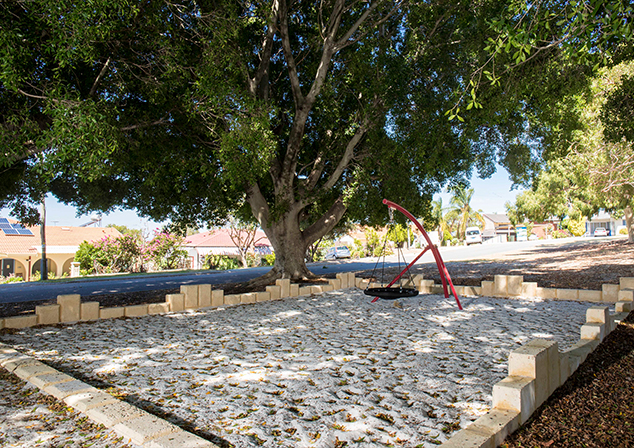 Image of large sandpit with a swing towards the back and a jagged limestone retaining wall. Large tree in background provides shade to the sandpit.