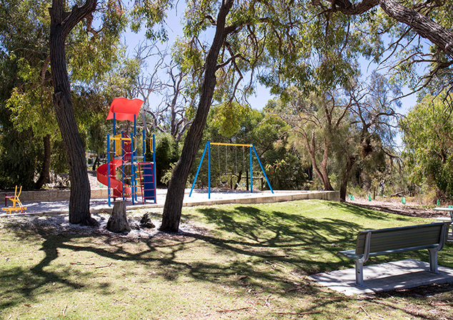 Picture of grass area with trees providing shade. Park bench in foreground, playground equipment in background