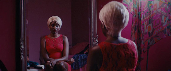 A woman in a red dress with a blonde wig stares into a mirror.
