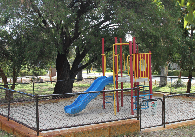 Image of sand pit with brightly coloured playground. Encased in a limestone wall with a chain link fence installed on top. Trees in background.