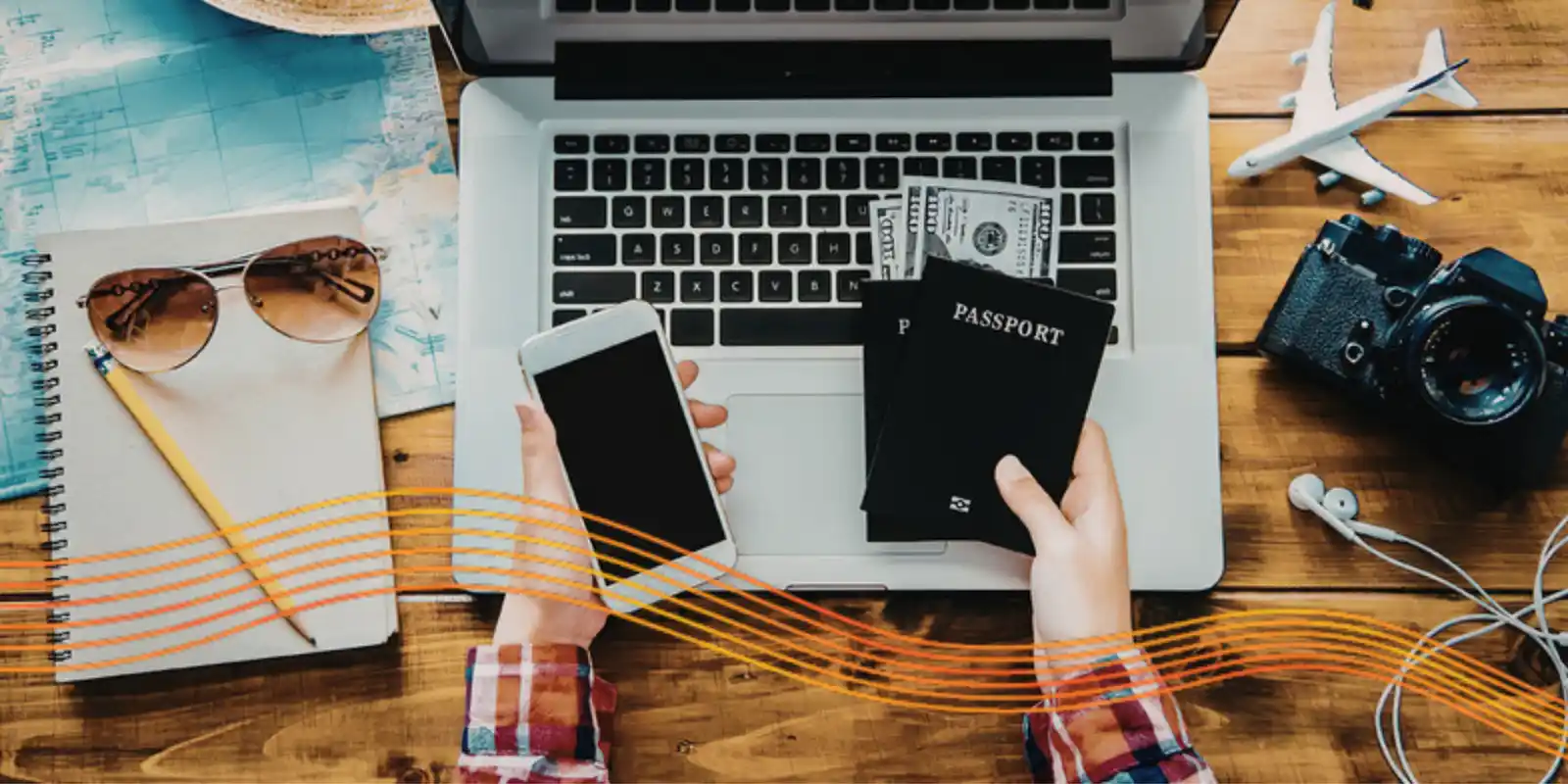 Two hands hold a smartphone, passports and cash over an open laptop. The laptop is surrounded by a map, notebook, sunglasses, pencil, camera, earbuds, and model aeroplane.