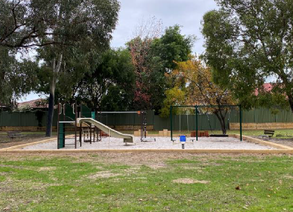 Photo of playground in sandpit, surrounded by grassy area with trees growing throughout. Fences in background.