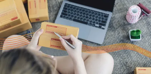 Someone writes an address onto a parcel. In the background is an open laptop.
